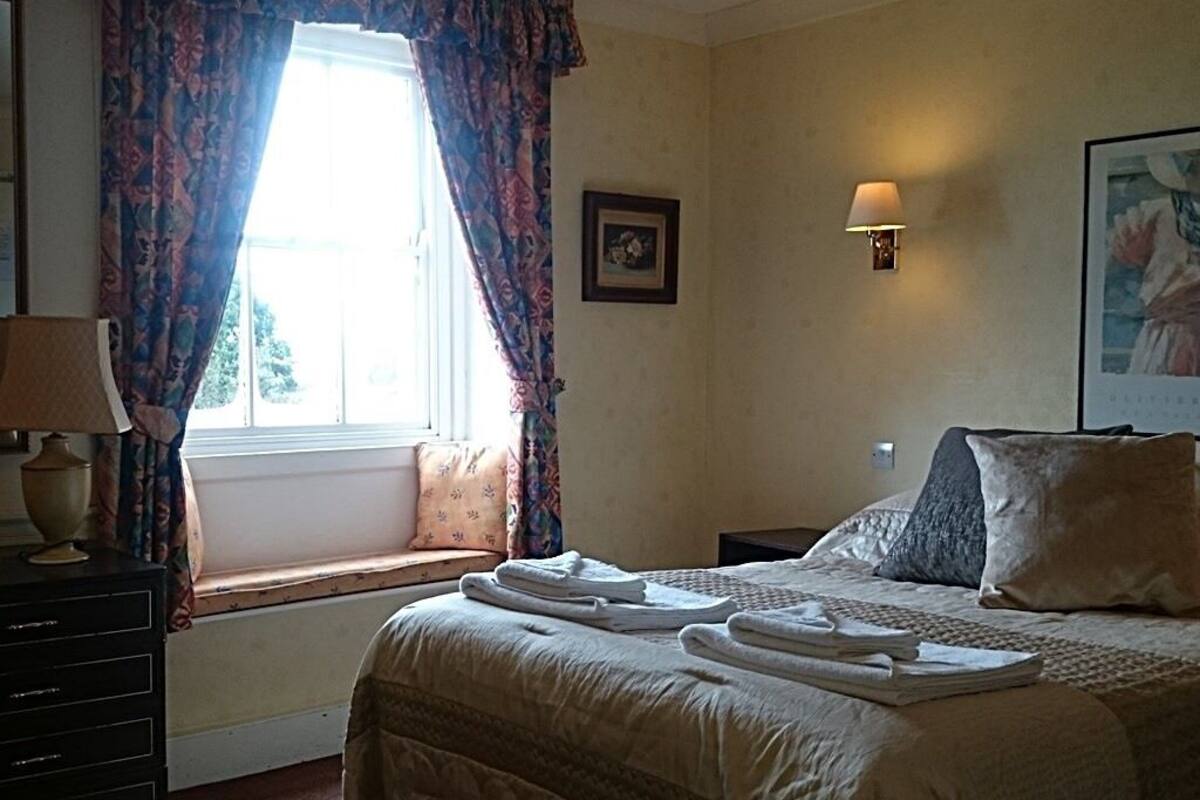 Una habitación de hotel con cama, mesita de noche, lámpara, ventana con cortinas y luz en la pared.