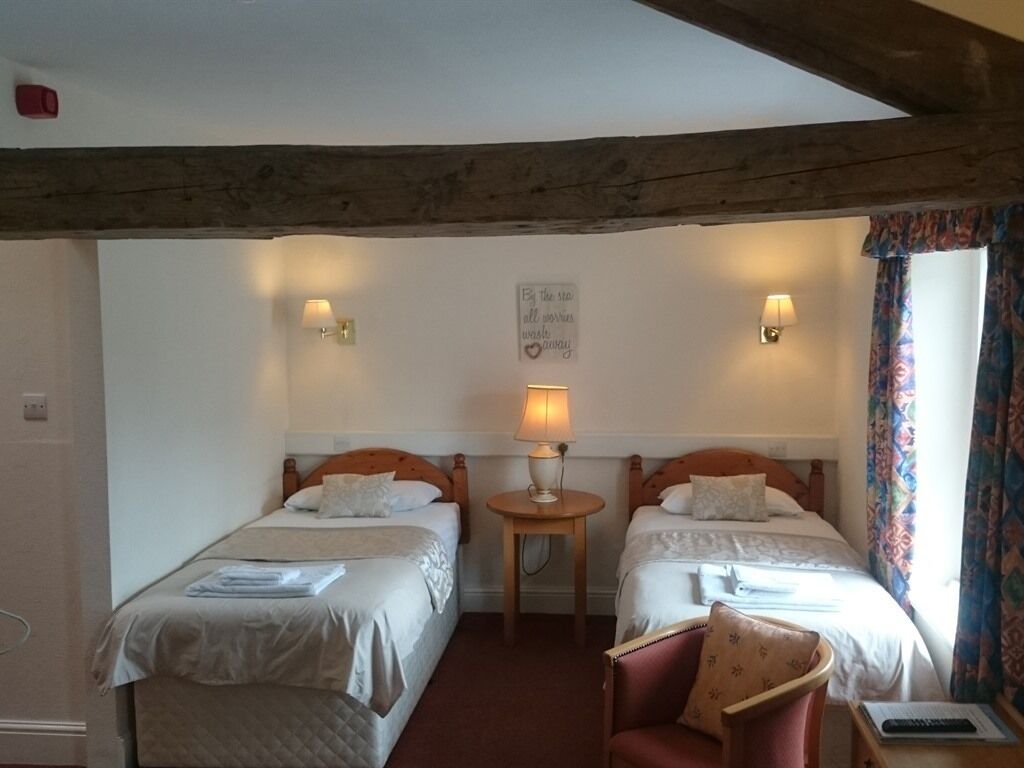 Habitación de hotel con dos camas individuales, una mesita con lámpara, una silla y una ventana con cortinas.
