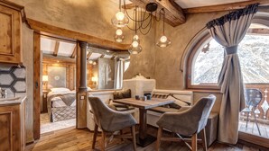 Un interior rústico con piso de madera, una zona de comedor con dos sillas, una mesa y una ventana con cortinas.