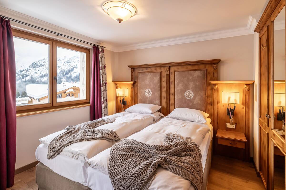A room with two beds, wooden headboard, bedside lamps, and a view of snow-capped mountains.