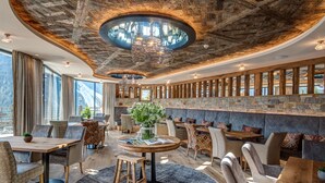 Un amplio y moderno salón con muebles de madera, detalles de piedra y grandes ventanales con vistas a las montañas.