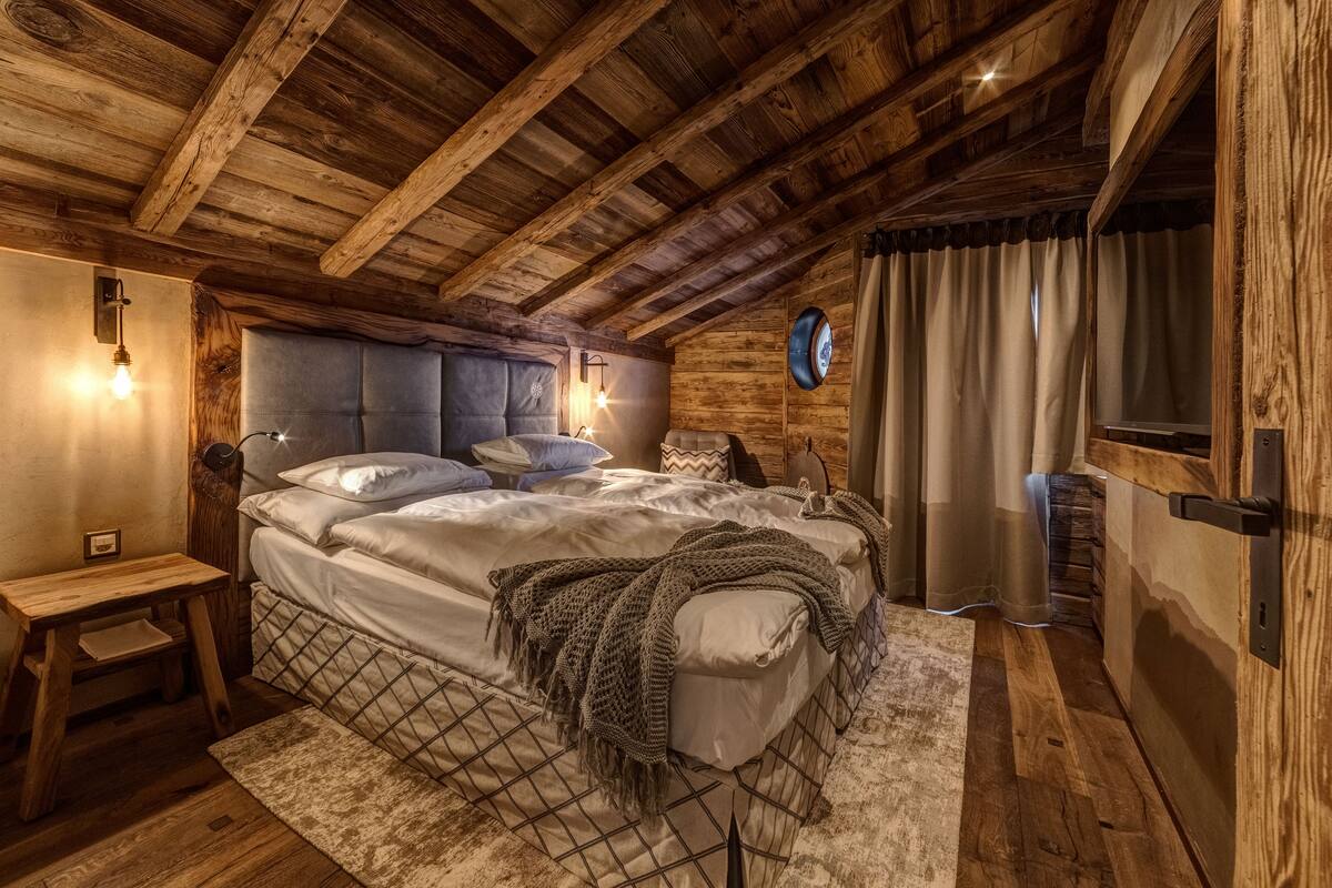 A cozy bedroom with a wooden ceiling, a bed with a knitted blanket, a bedside table, and a wall-mounted clock.