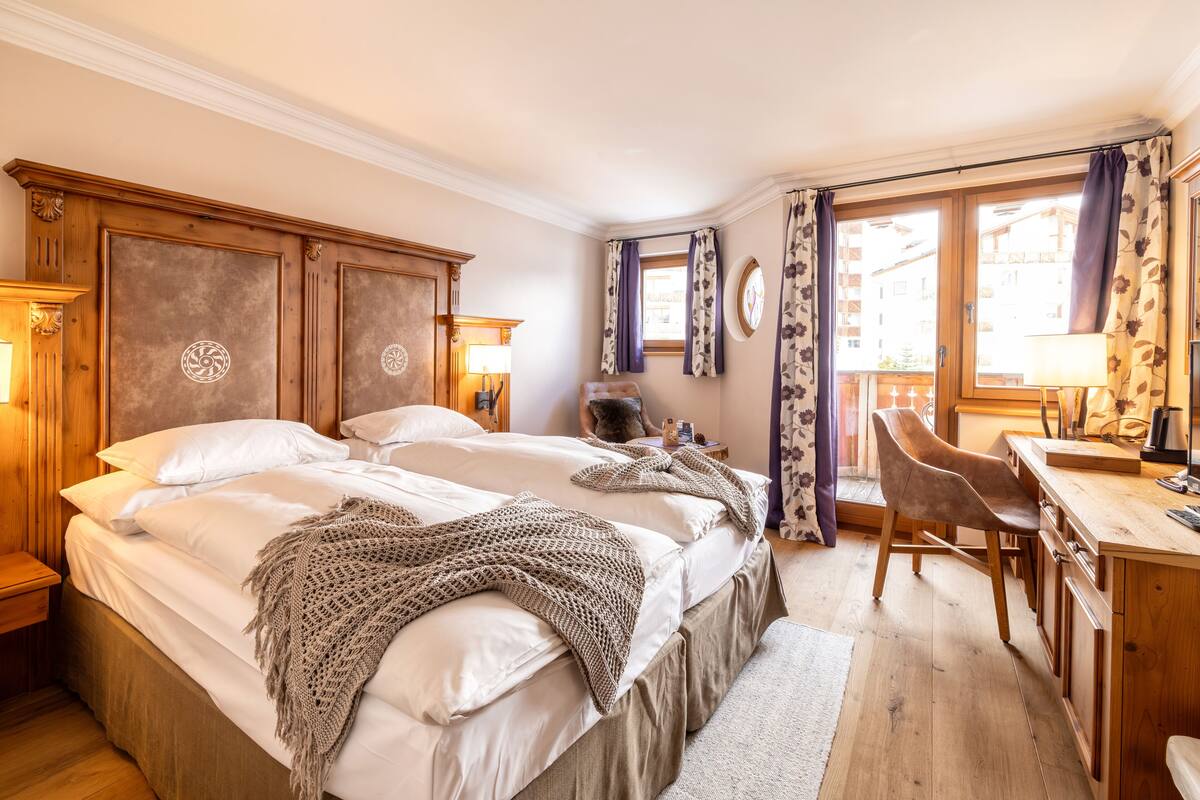 A hotel room with a large bed, a wooden desk, and a chair. There is a window with curtains, a lamp, and a clock on the wall.