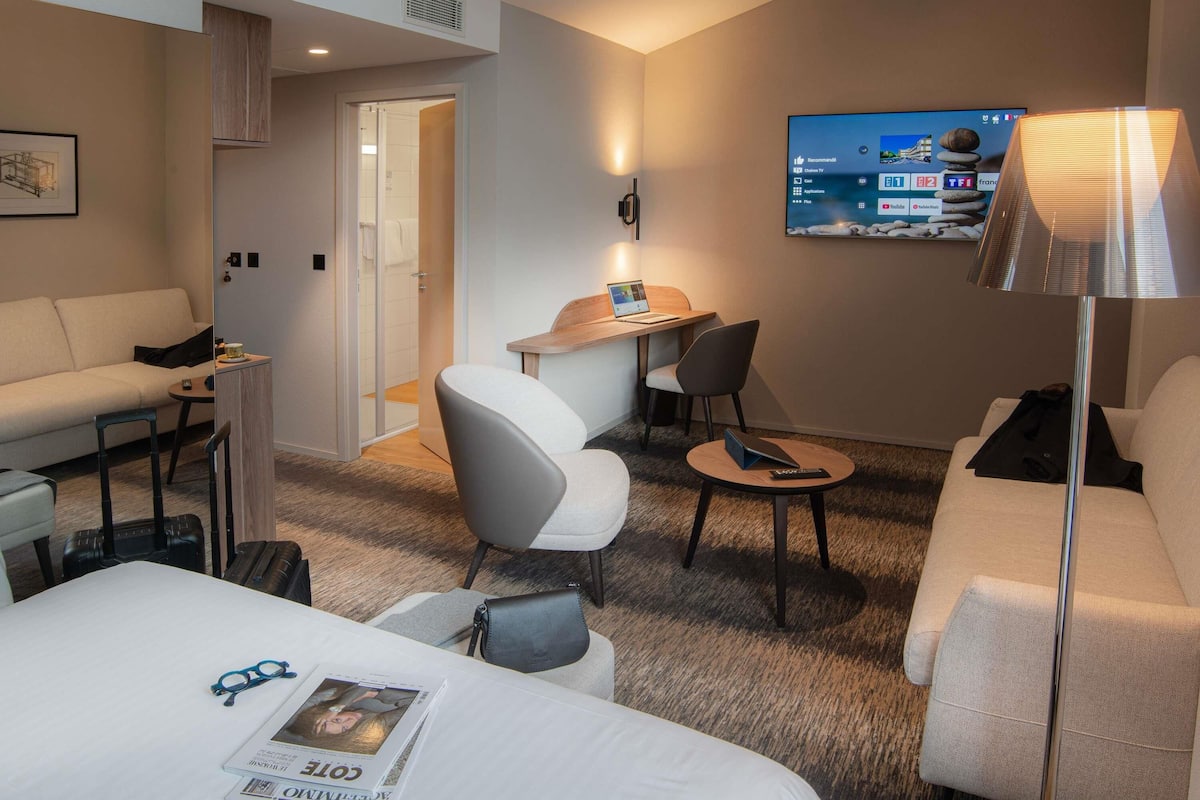 Une chambre d’hôtel avec un lit, un bureau, une chaise, un canapé et un téléviseur fixé au mur.