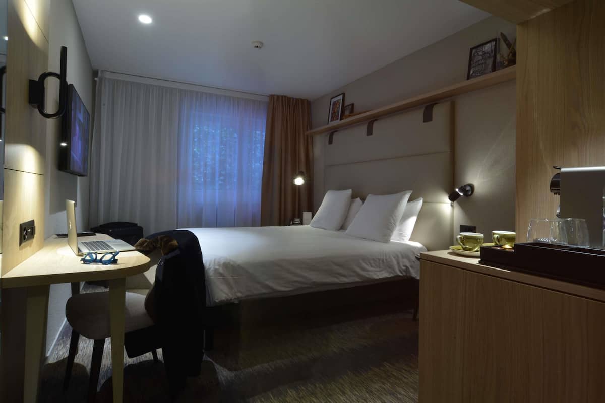 Une chambre d’hôtel équipée d’un lit, d’un bureau avec un ordinateur portable, d’une chaise, d’une machine à café et d’un téléviseur fixé au mur.