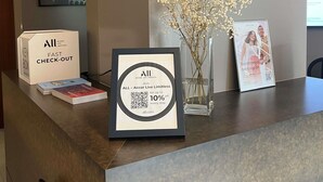 Una recepción de hotel con un cartel enmarcado que ofrece un descuento del 10% por hospedarse en el hotel, una caja de llaves, una foto enmarcada y un jarrón con flores secas.