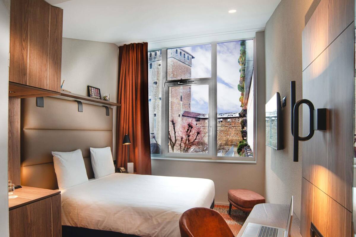 Une chambre d’hôtel avec un lit, un bureau, un ordinateur portable et une vue sur un immeuble par la fenêtre.