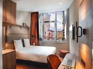 A hotel room with a bed, desk, laptop, and a view of a building through the window. - Mercure Fribourg Centre Remparts (Fribourg)