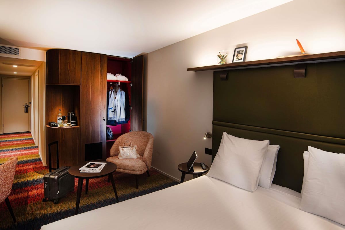 Une chambre d’hôtel avec un grand lit, un petit fauteuil, un bureau avec une lampe, une armoire et une moquette colorée.