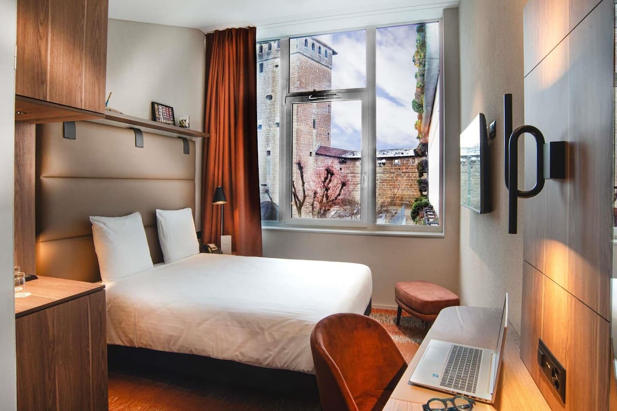 A modern hotel room with a bed, desk, laptop, and a large window with a city view.