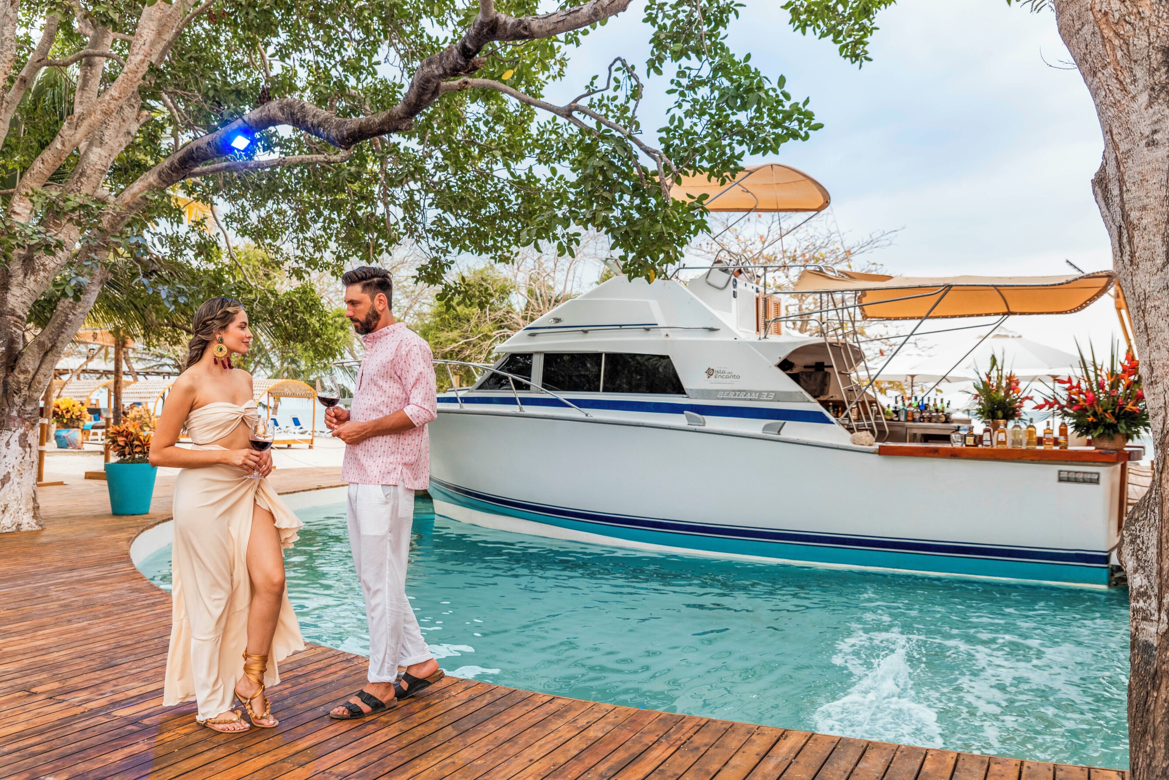 A couple standing by a yacht docked at a poolside area with a wooden deck and potted plants.