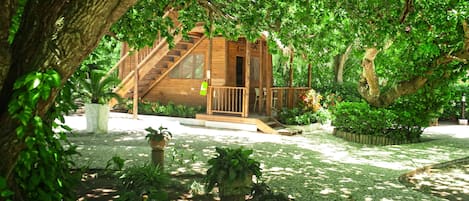 A wooden cabin with a staircase leading to a deck, surrounded by lush greenery and a cobblestone path.