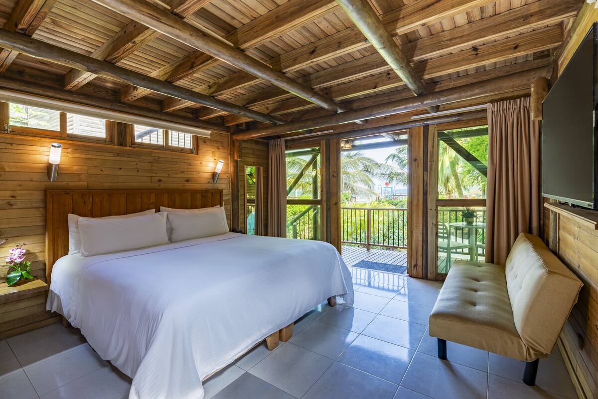 A wooden cabin room with a bed, a bench, and large windows overlooking a garden.
