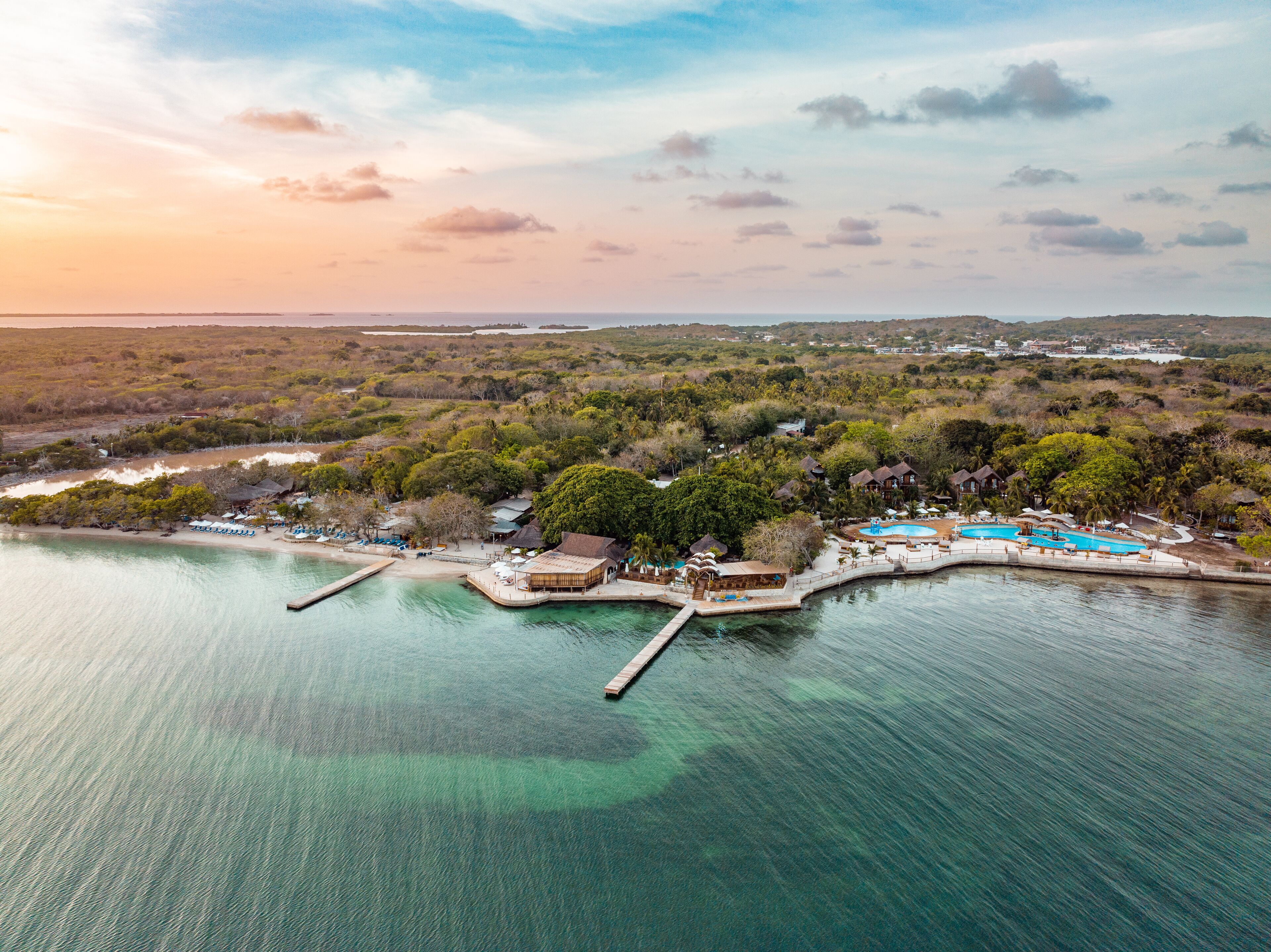 An aerial view of a coastal resort with multiple buildings, a swimming pool, and a dock extending into the water.