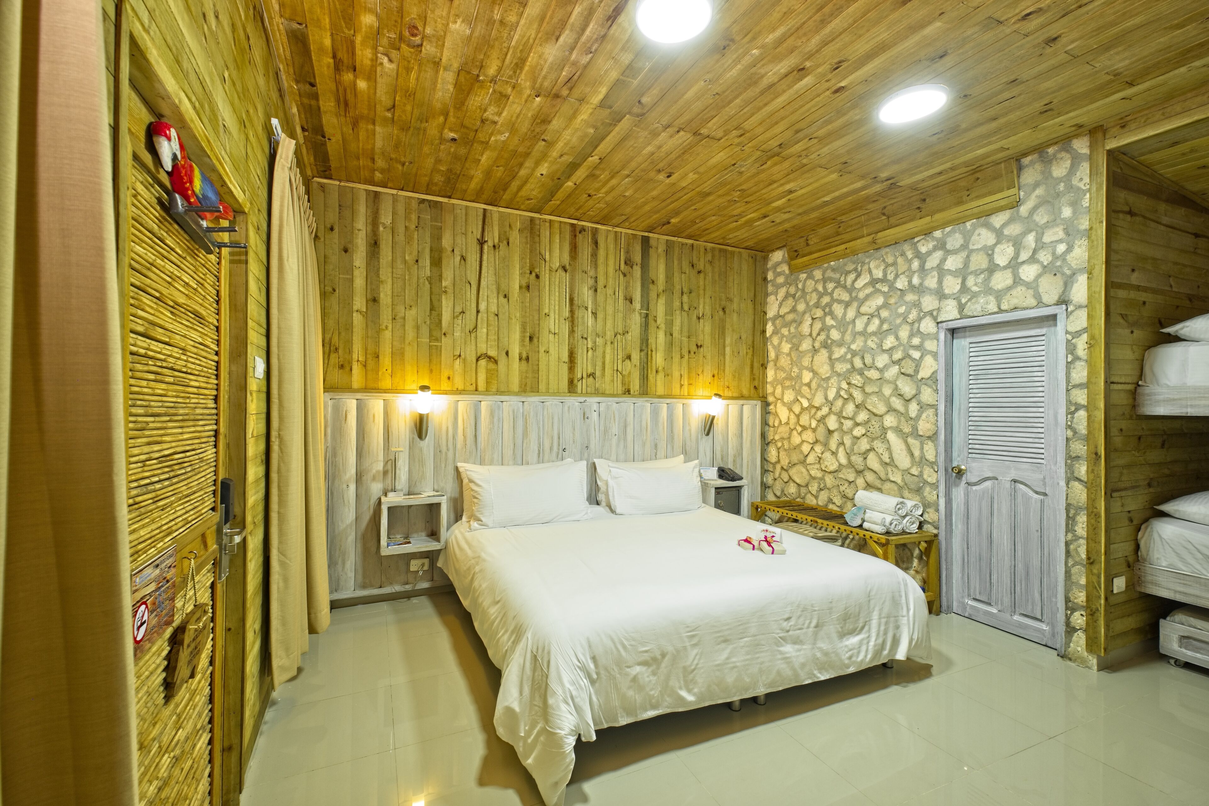 Una habitación con techo de madera, pared de piedra, cama con ropa blanca, mesita de noche, lámpara y armario.