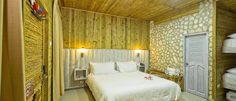 Una habitación con techo de madera, pared de piedra, cama con ropa blanca, mesita de noche, lámpara y armario.