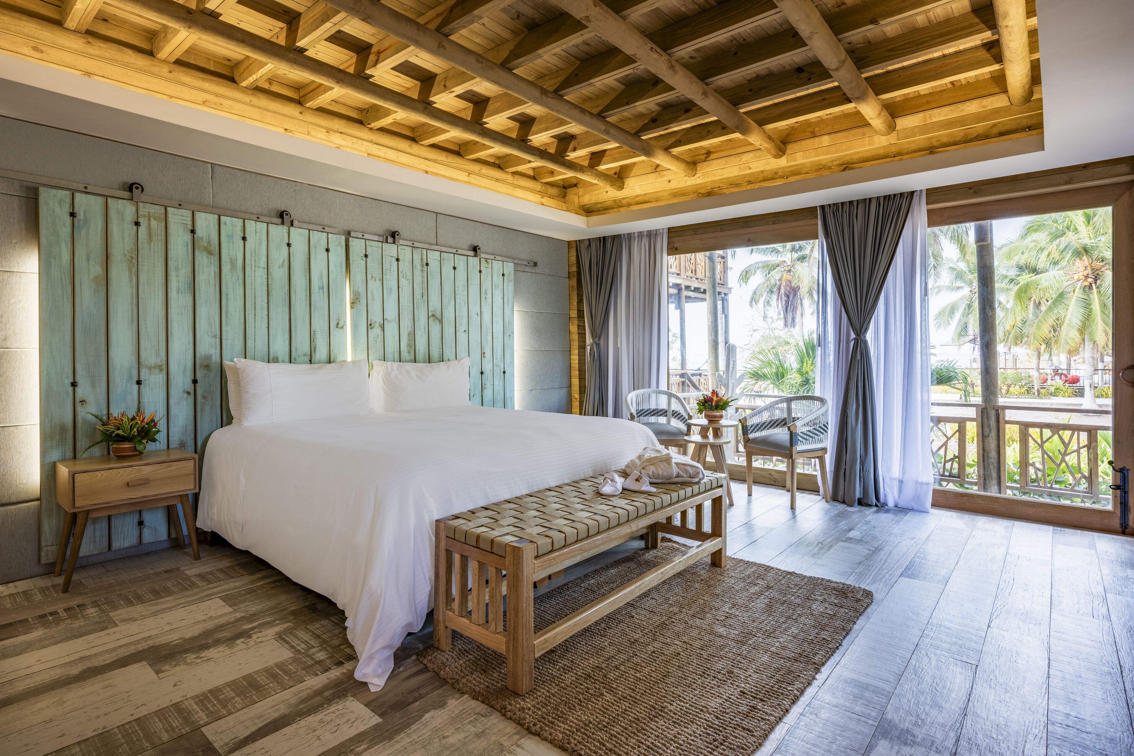 A bedroom with a wooden ceiling, a large bed, a bench, a nightstand, and a view of palm trees outside.