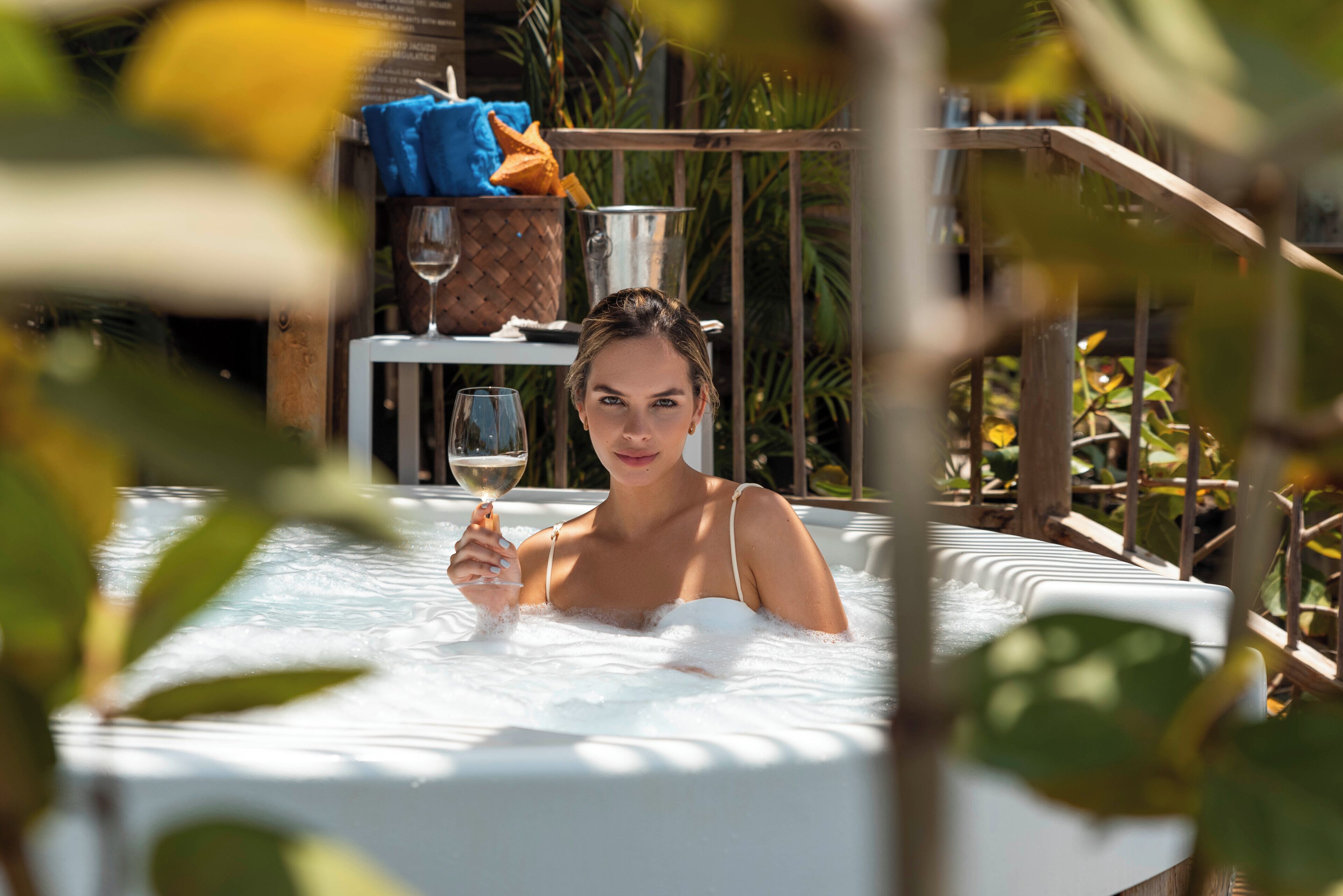 A person in a hot tub with a glass of wine, surrounded by greenery.