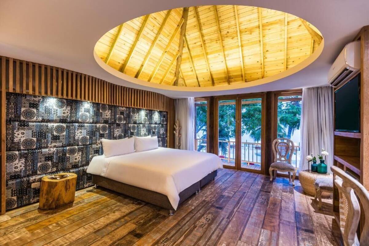 A bedroom with a large bed, a wooden ceiling, a patterned wall, and a view of the outdoors.
