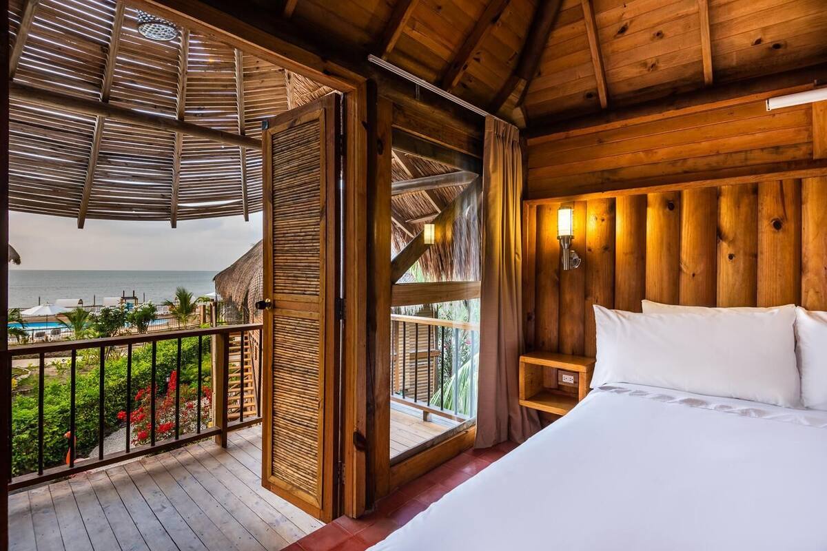 A wooden room with a bed, a balcony with a view of the sea, and a thatched roof.