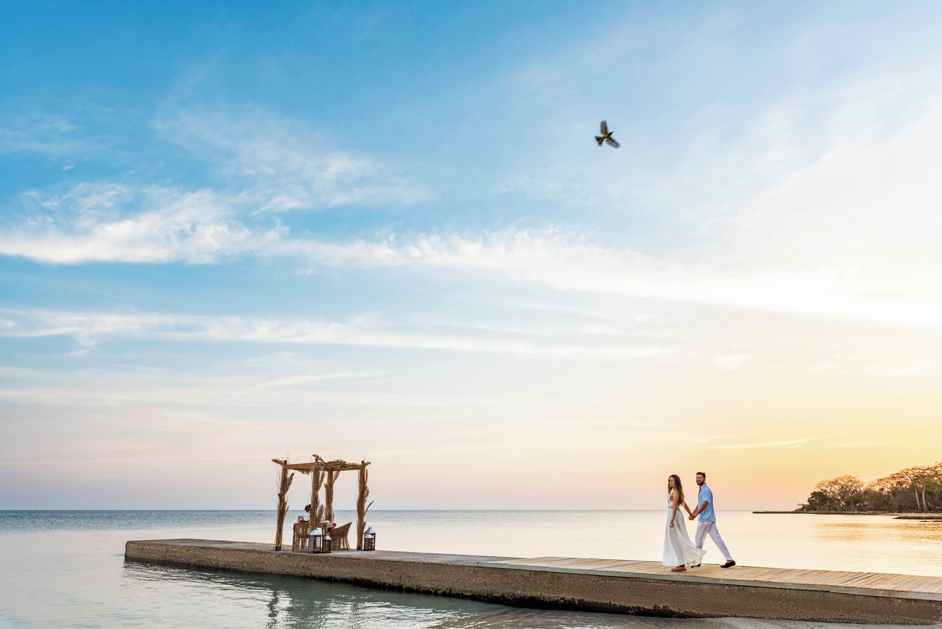 Una pareja caminando sobre un muelle con una estructura de madera y un ave volando en el cielo.