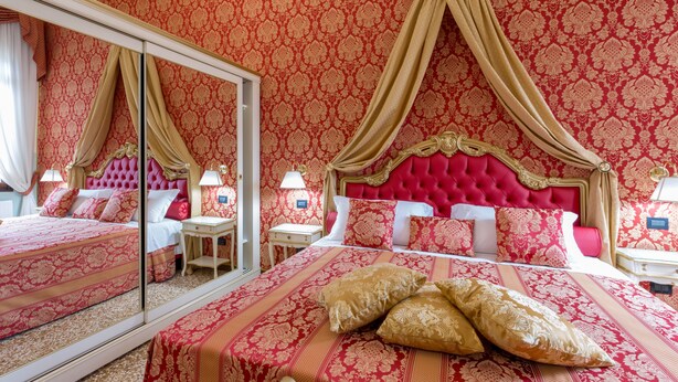 A luxurious bedroom with a large mirror, ornate red and gold wallpaper, a tufted red headboard, and a neatly made bed with matching pillows.