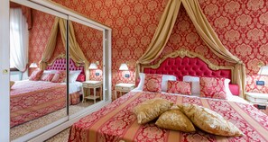 A luxurious bedroom with a large mirror, ornate red and gold wallpaper, a tufted red headboard, and a neatly made bed with matching pillows. - Friendly Venice Suites (Venice)