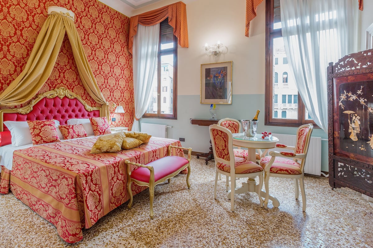 A room with a large bed, a sitting area with a table and chairs, and a view of buildings outside the window.