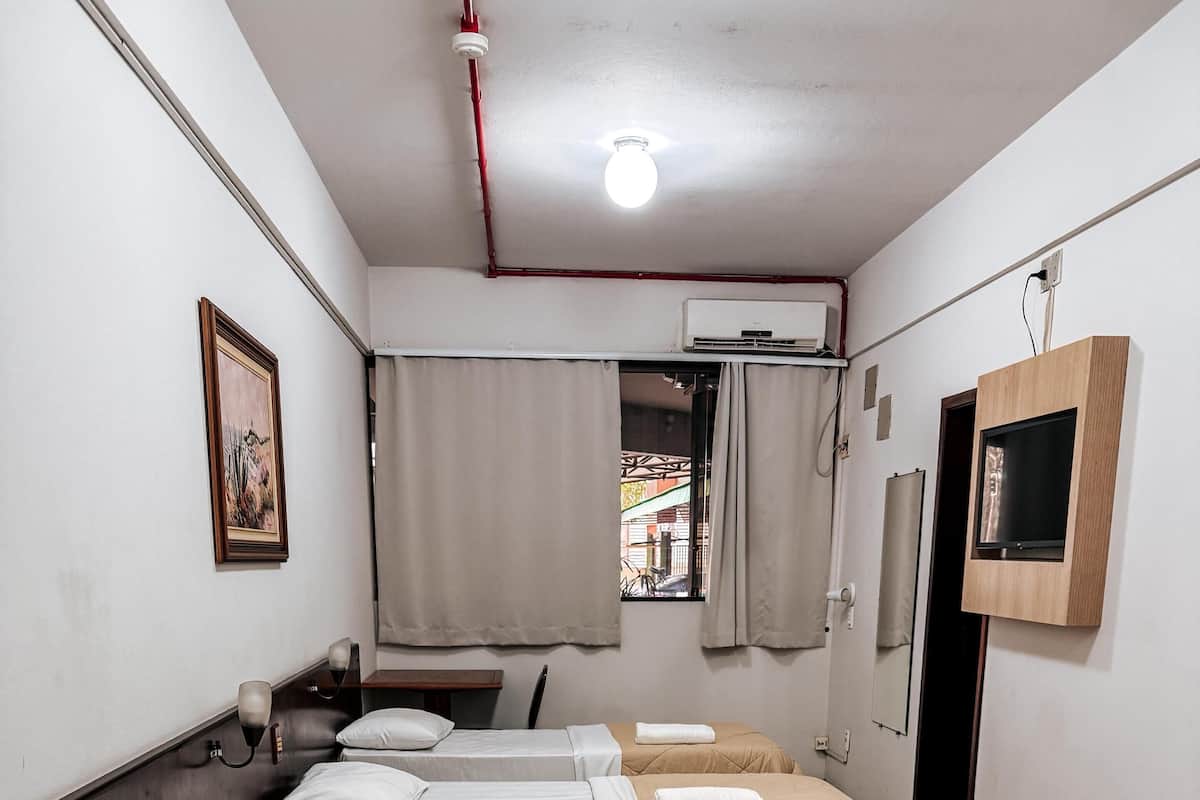 A hotel room with three beds, a window with curtains, a mounted TV, and a painting on the wall.