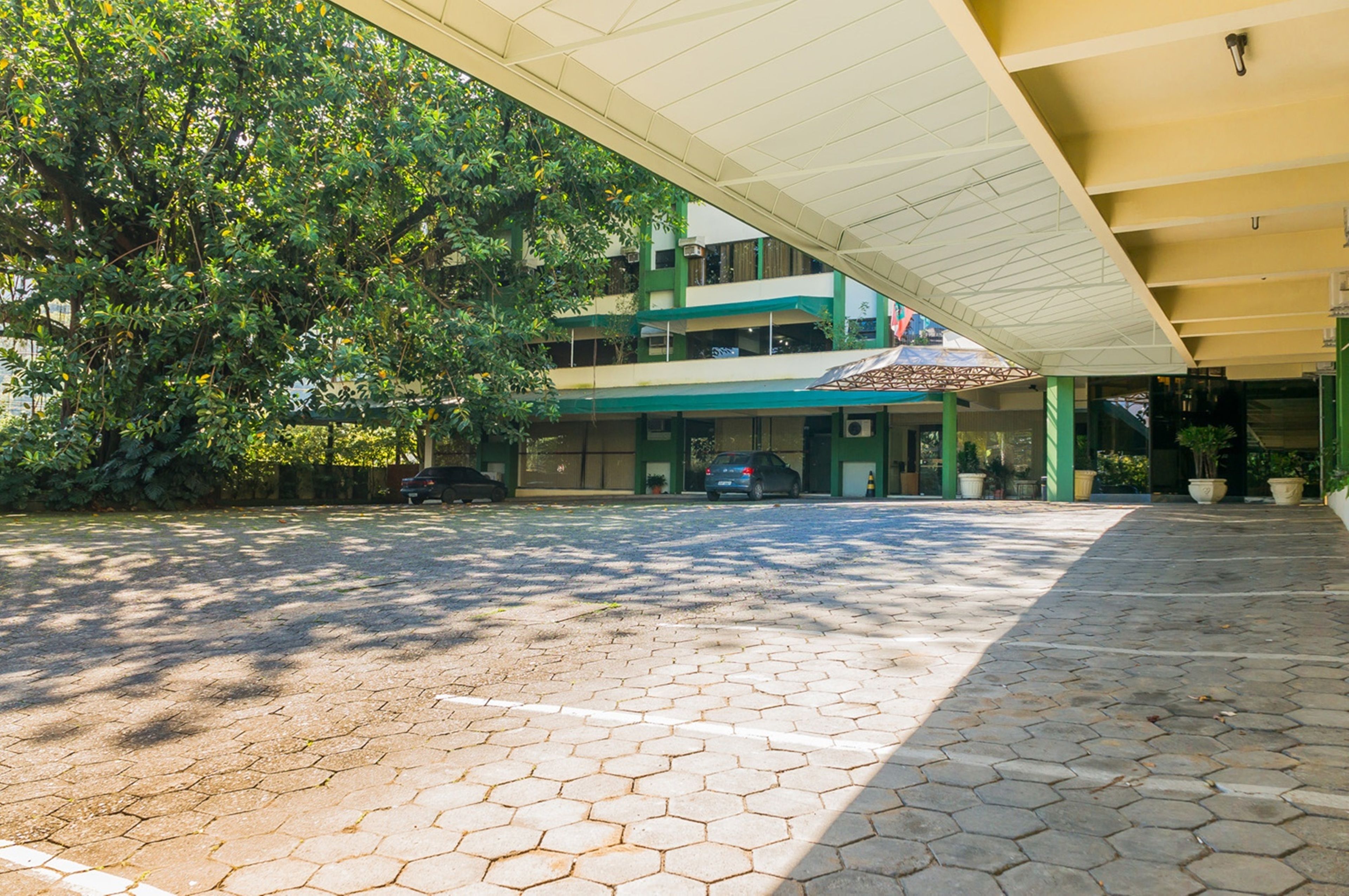 A covered outdoor area with a paved walkway, a building with multiple floors, and a large tree providing shade.