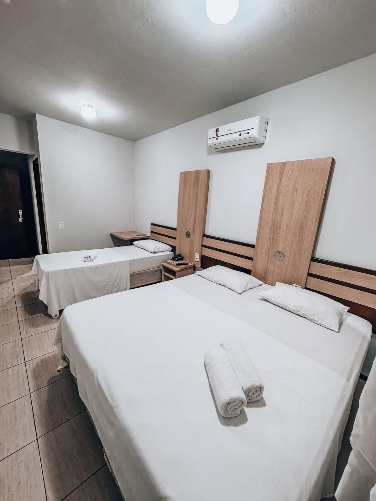 A hotel room with two beds, a bedside table, a nightstand, and an air conditioning unit on the wall.