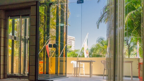 A modern building entrance with glass doors, a small outdoor seating area, and a view of palm trees and a flag. - Harbor Inn Jaraguá (Jaragua do Sul)