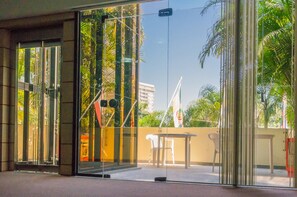 A modern building entrance with glass doors, a small outdoor seating area, and a view of palm trees and a flag. - Harbor Inn Jaraguá (Jaragua do Sul)