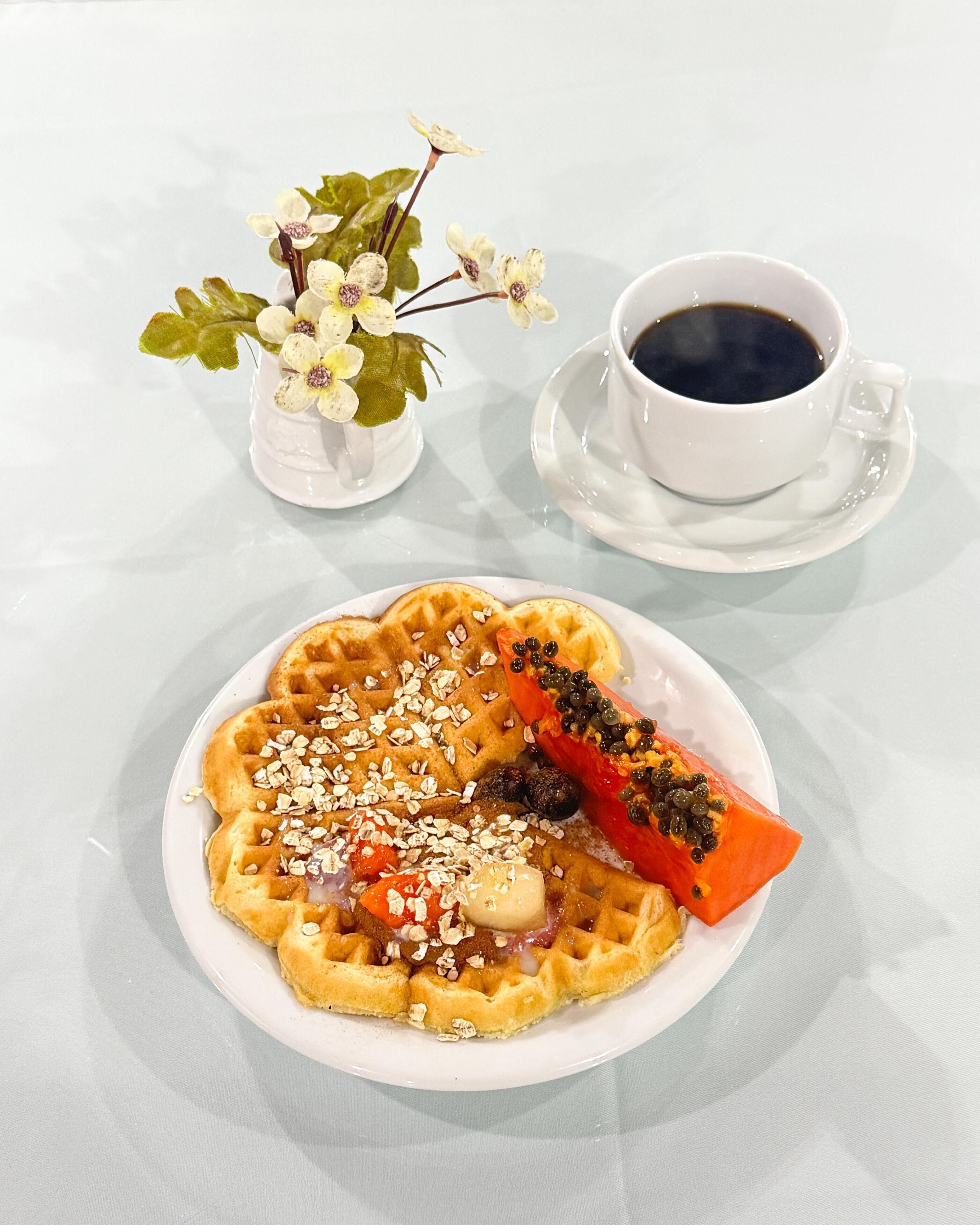 Un plato de gofres con acompañamientos, una taza de café y un pequeño jarrón con flores sobre una mesa.