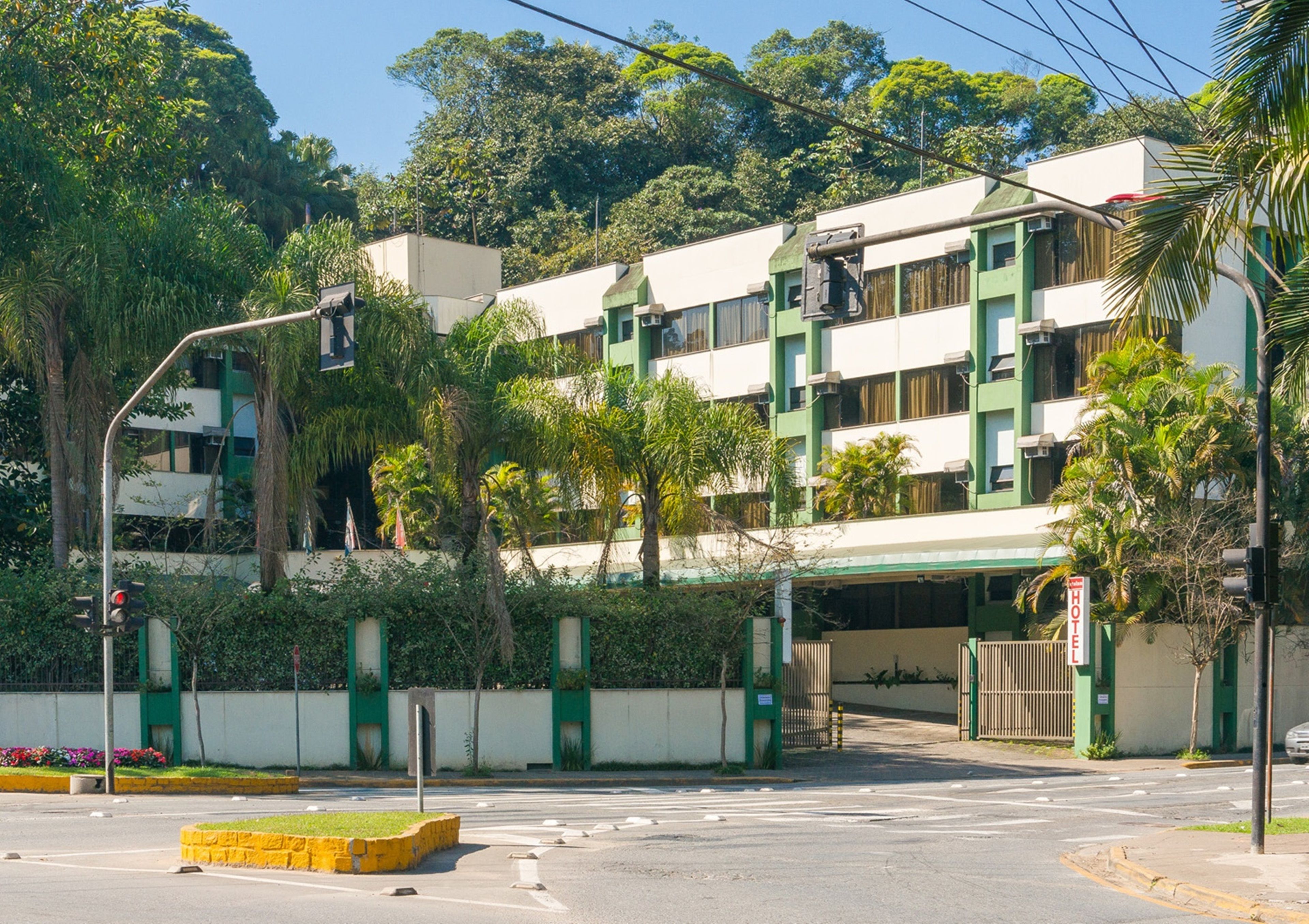 A multi-story hotel with a green and white facade, a gated entrance, and a parking area with yellow curbs.