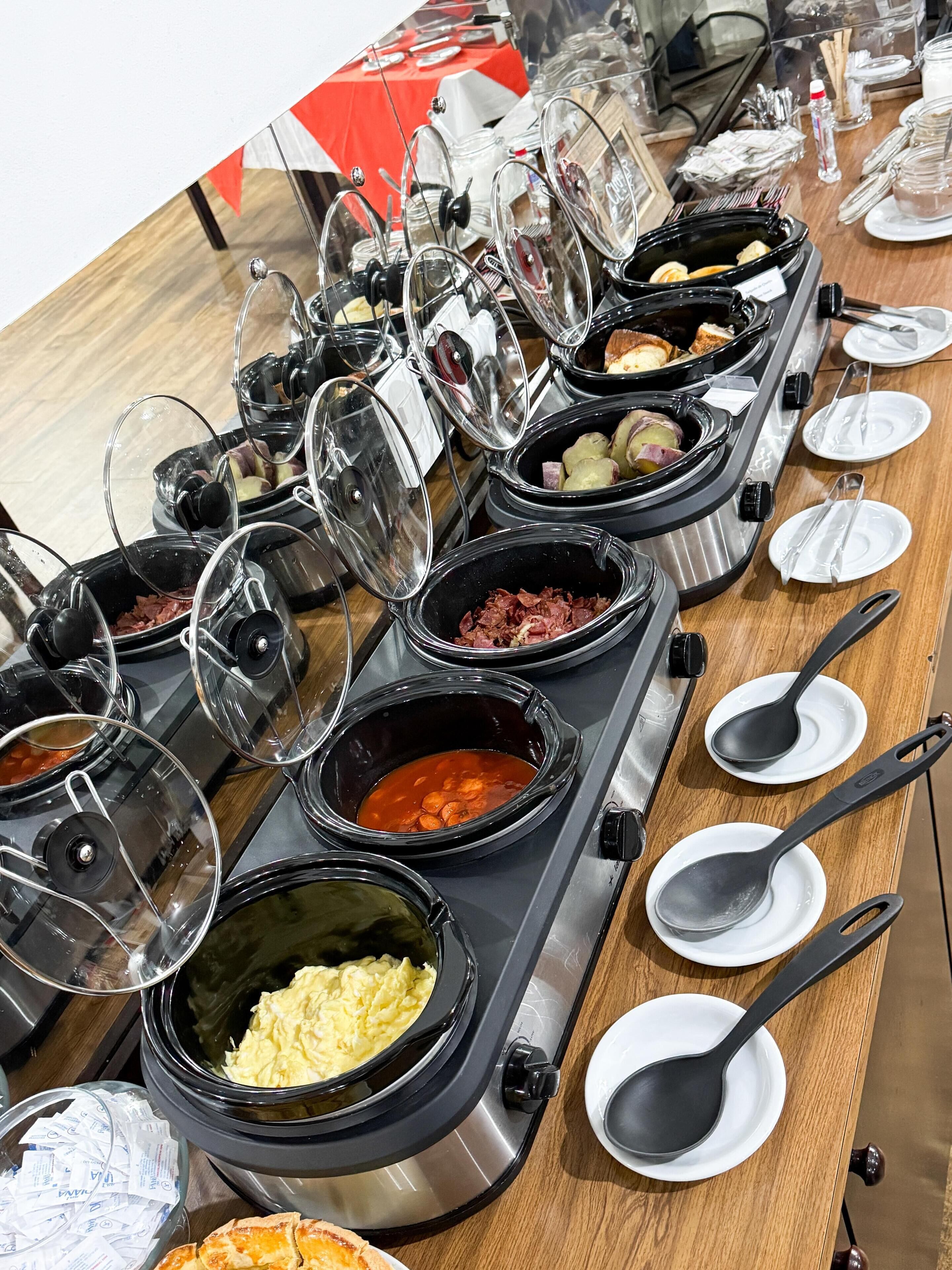 Disposición de un bufé con diferentes alimentos en recipientes cubiertos, colocados sobre una mesa con platos y cucharas.