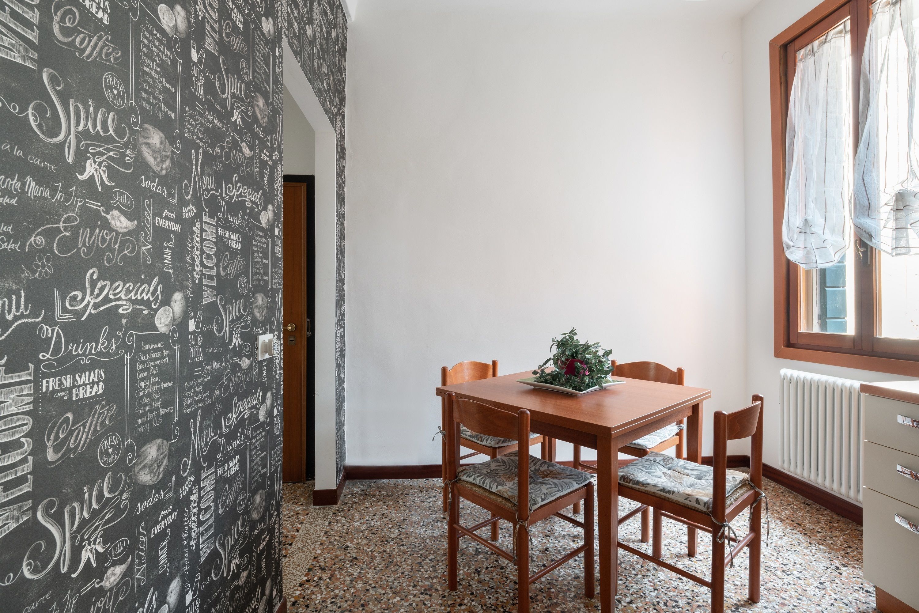 Ein Essbereich mit einem Holztisch und Stühlen, einer Zierpflanze und einer Wand mit verschiedenen Wörtern rund um Kaffee.