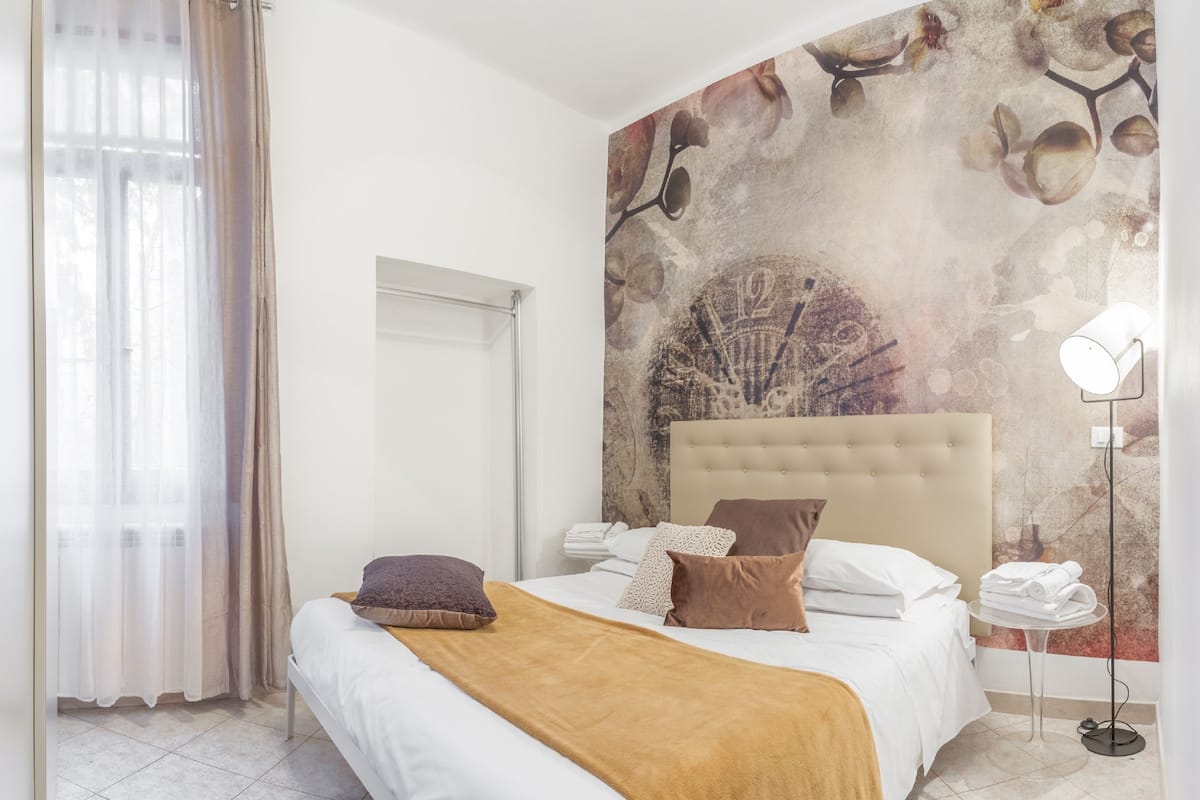 A bedroom with a bed, a headboard, a wall mural, a window with curtains, and a bedside table with a lamp.