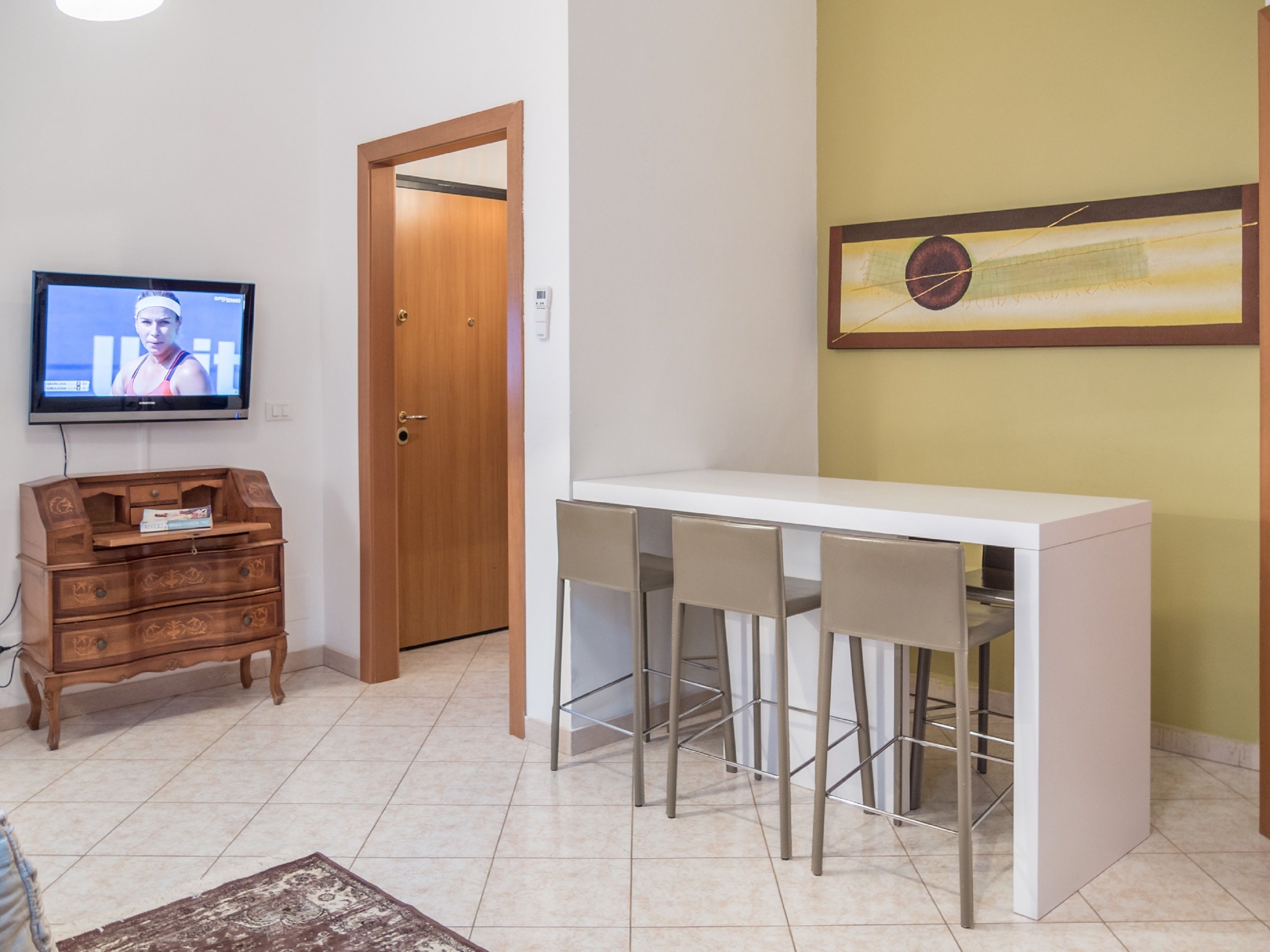A compact living space with a white dining table and two chairs, a wooden dresser, a television, and a door leading to another room.