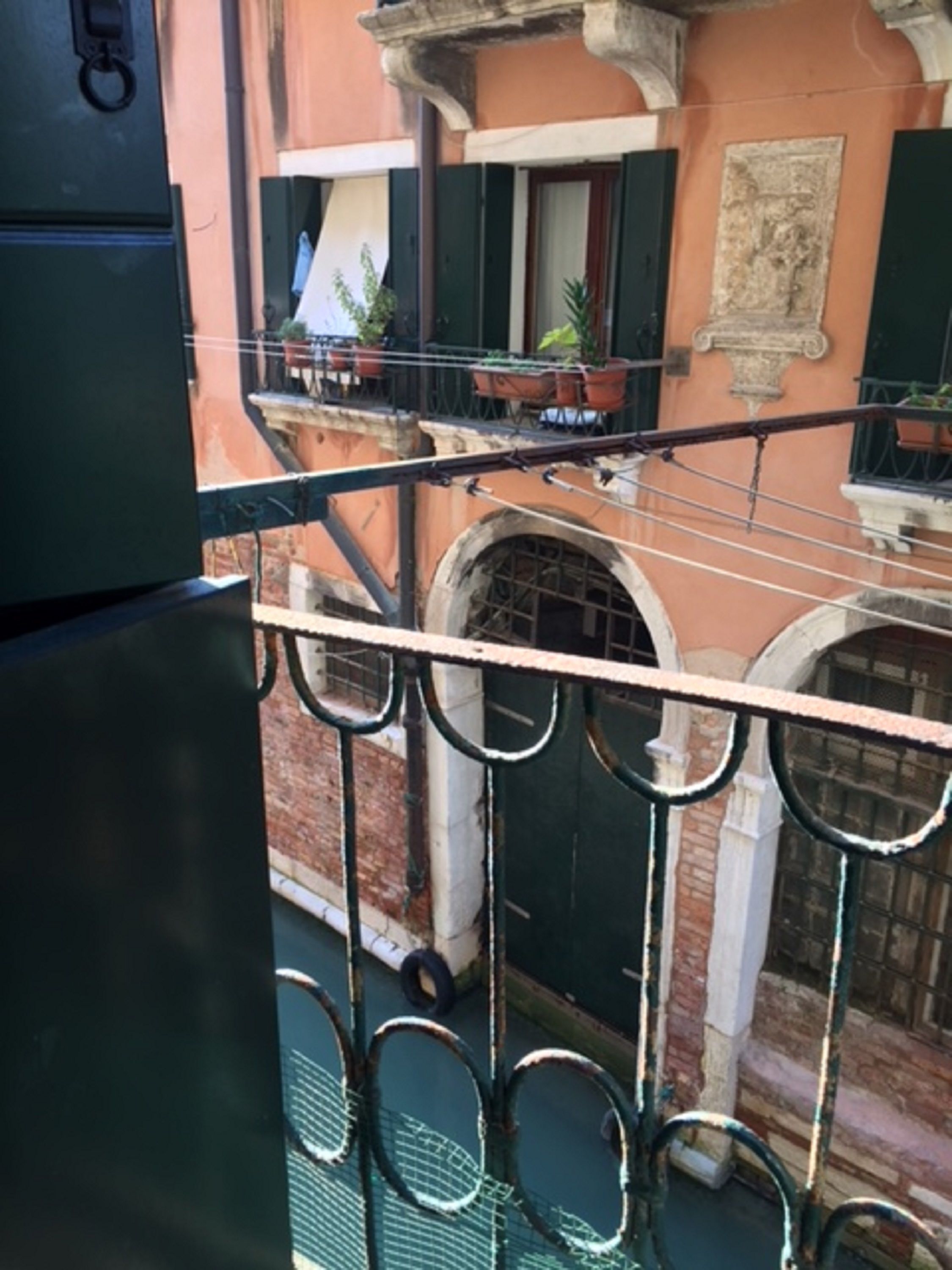 A balcony with a metal railing, a view of a building with a stone facade, and a window with a green shutter.