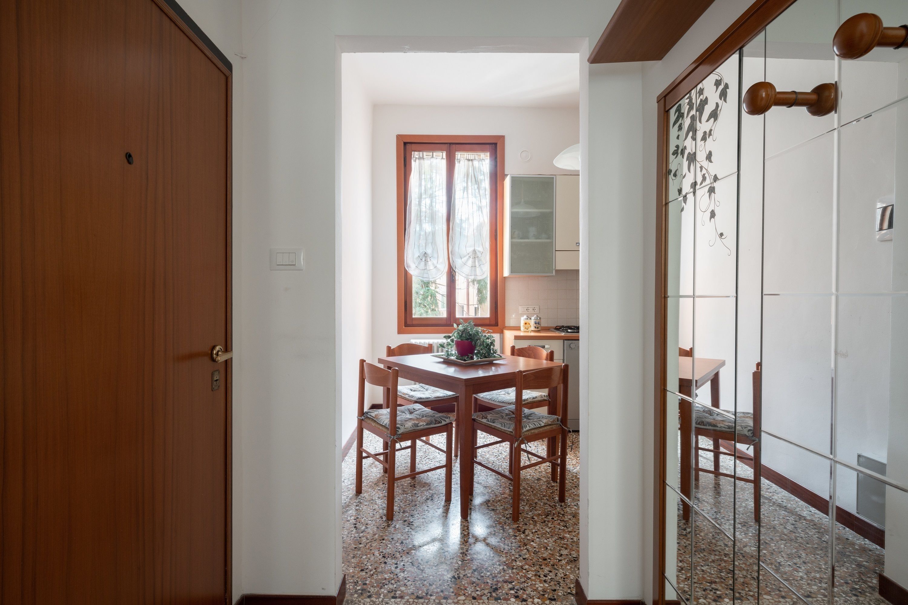 A dining area with a wooden table and chairs, a window with curtains, and a kitchen in the background.