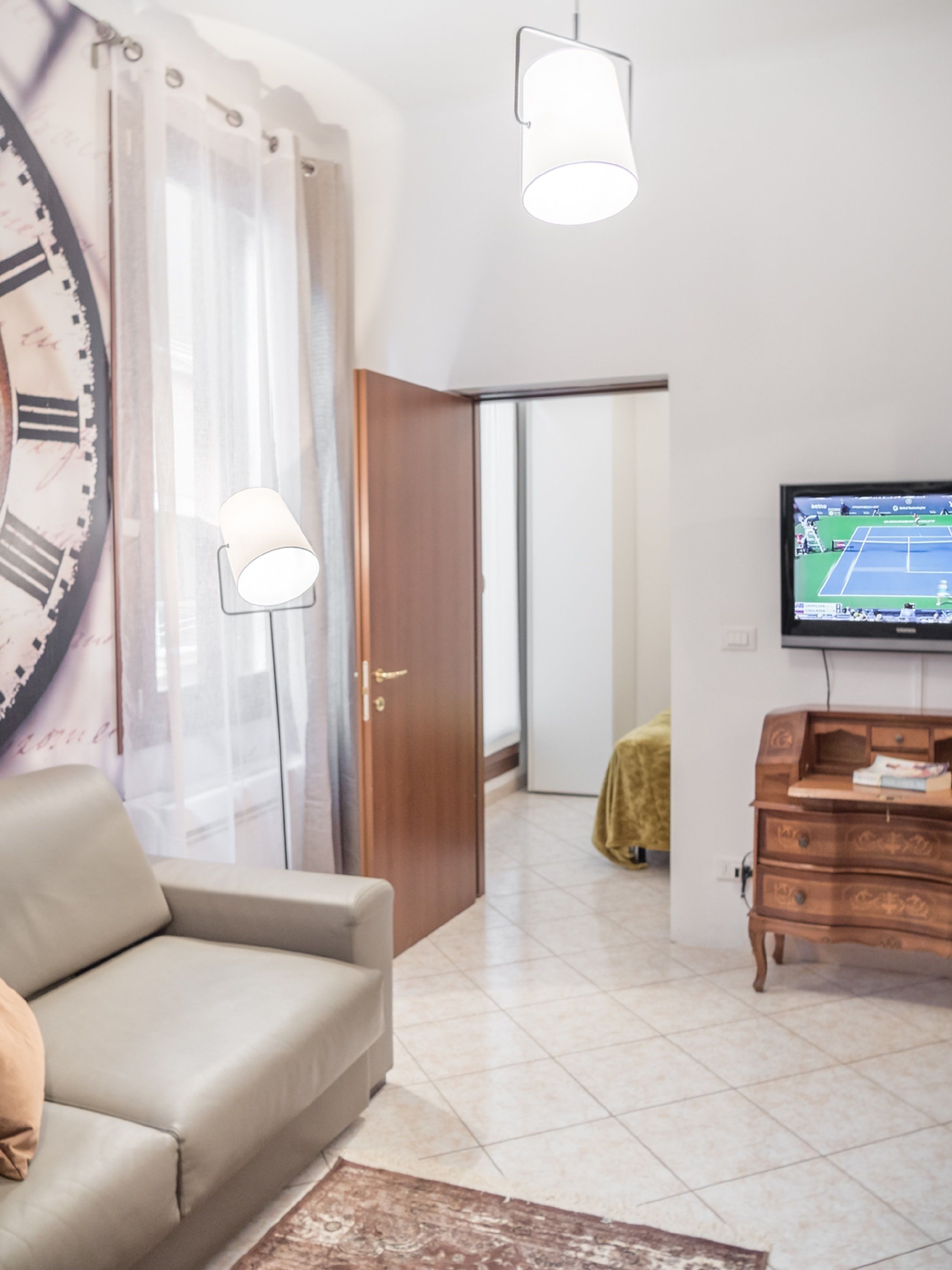 Een moderne woonkamer met een bank, een televisie waarop een tenniswedstrijd te zien is, een houten dressoir en een grote wandklok.
