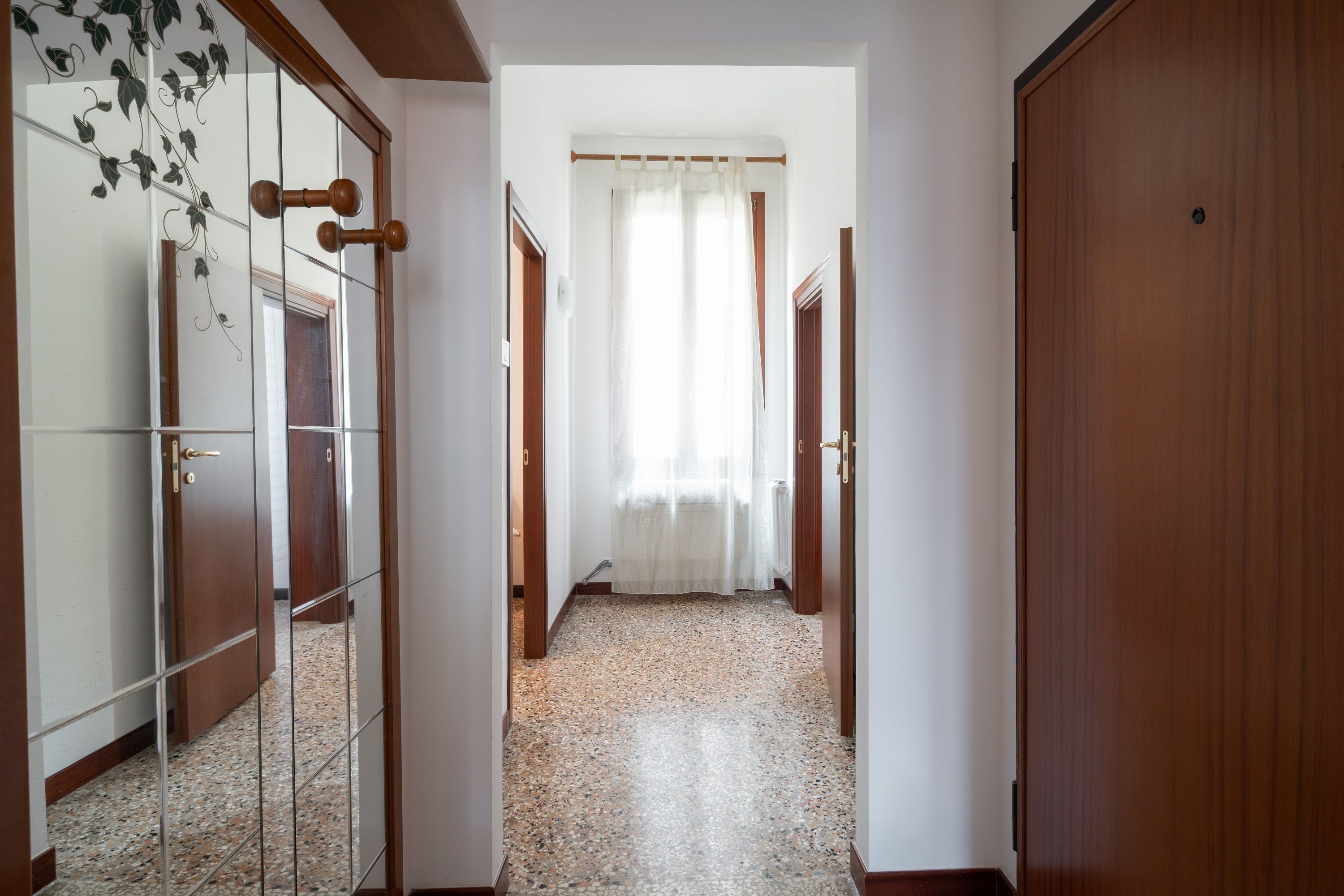 A hallway with a tiled floor, wooden doors, and a window with sheer curtains.