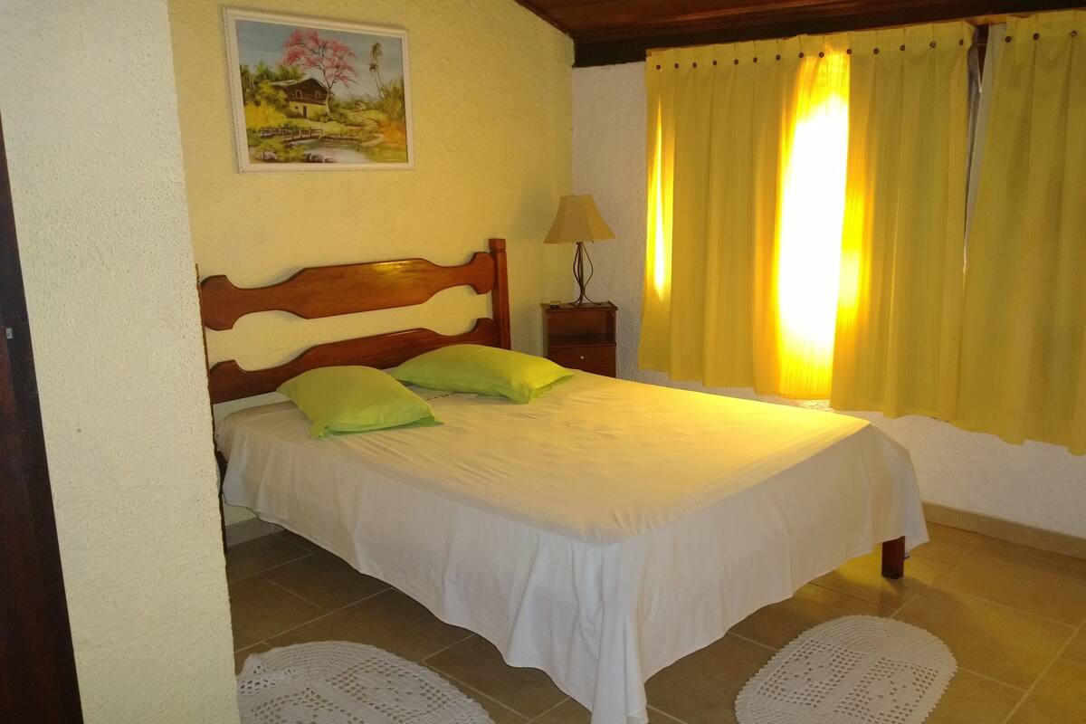 A bedroom with a wooden headboard, a bed with white bedding, two green pillows, a nightstand with a lamp, and a painting on the wall.