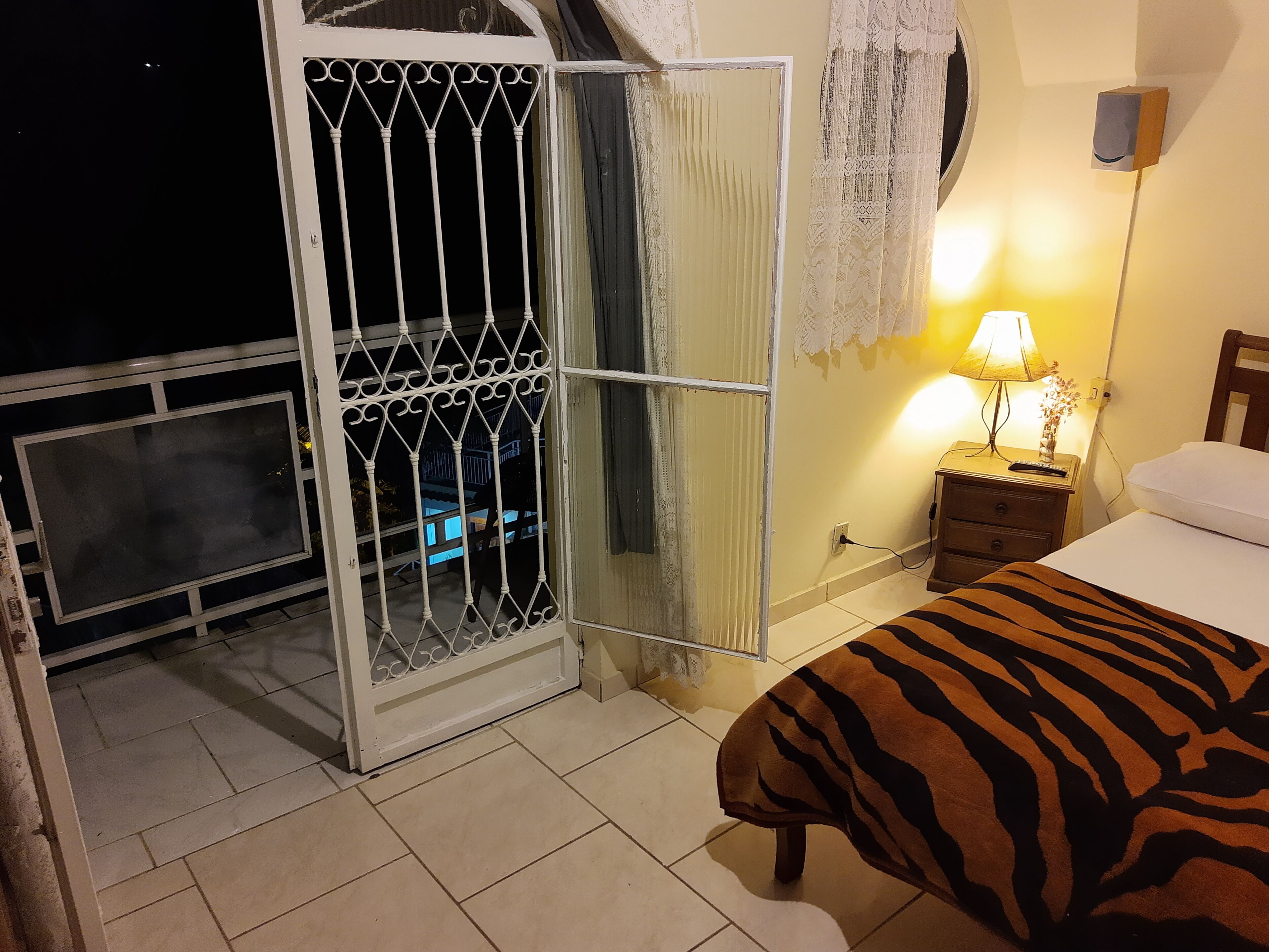 Um quarto com uma cama, mesa de cabeceira, abajur, janela com cortinas de renda e varanda com grade de metal.