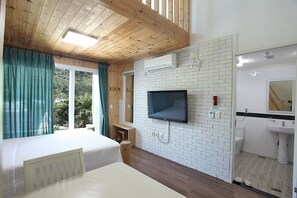 A room with a bed, a TV mounted on a brick wall, a window with curtains, and a bathroom with a sink and mirror. - Mirvill Pension (Seogwipo)
