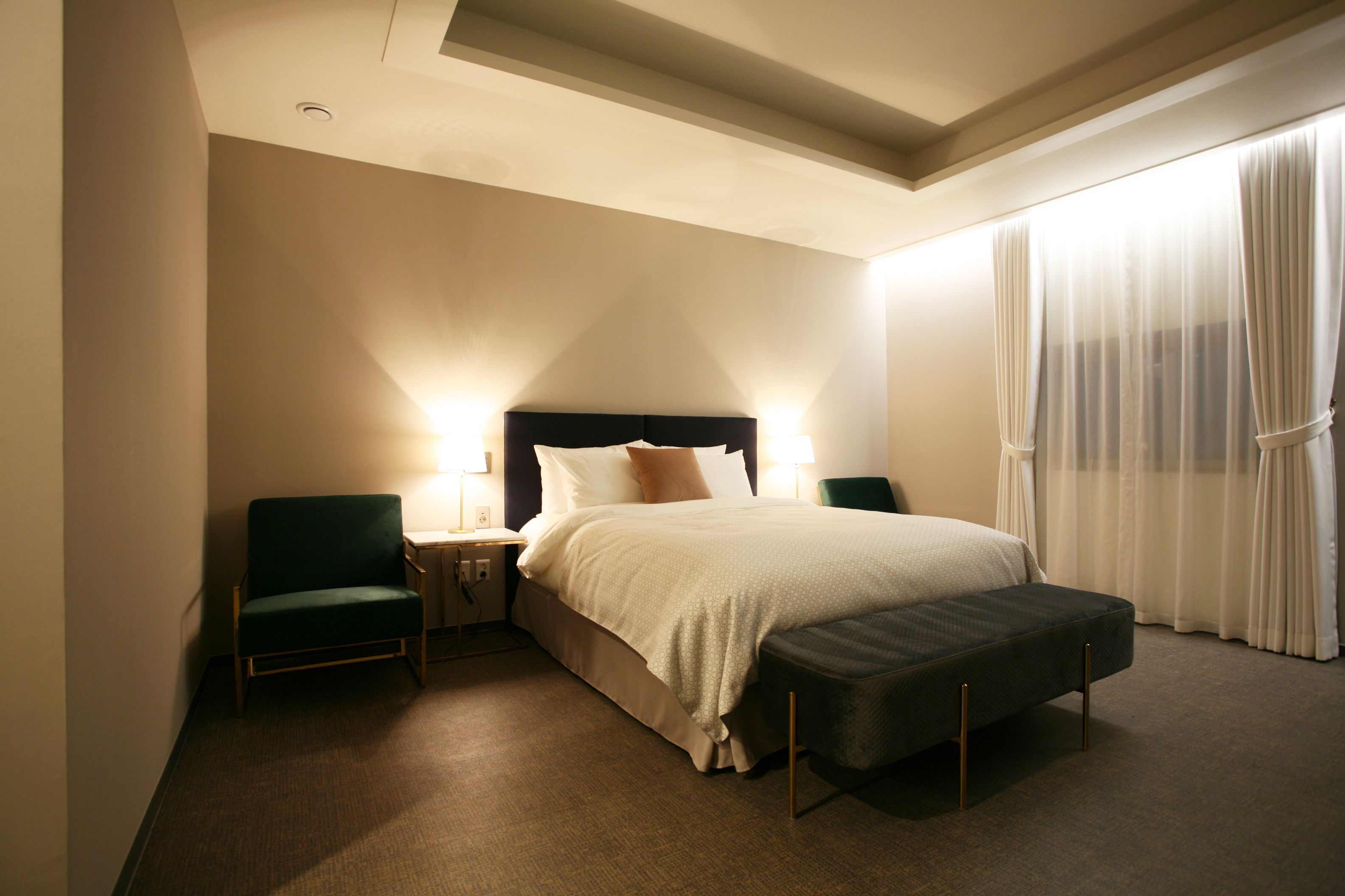 A hotel room with a bed, two nightstands, a chair, a bench, and a large window with curtains.