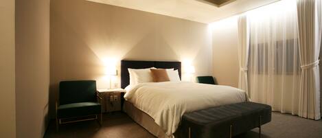 Quarto de hotel com uma cama, duas mesas de cabeceira, uma cadeira, um banco e uma janela grande com cortinas.