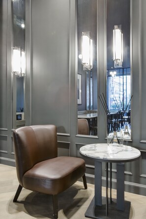 A modern hotel lobby with a leather armchair, a marble table, and a view of the dining area through large windows.