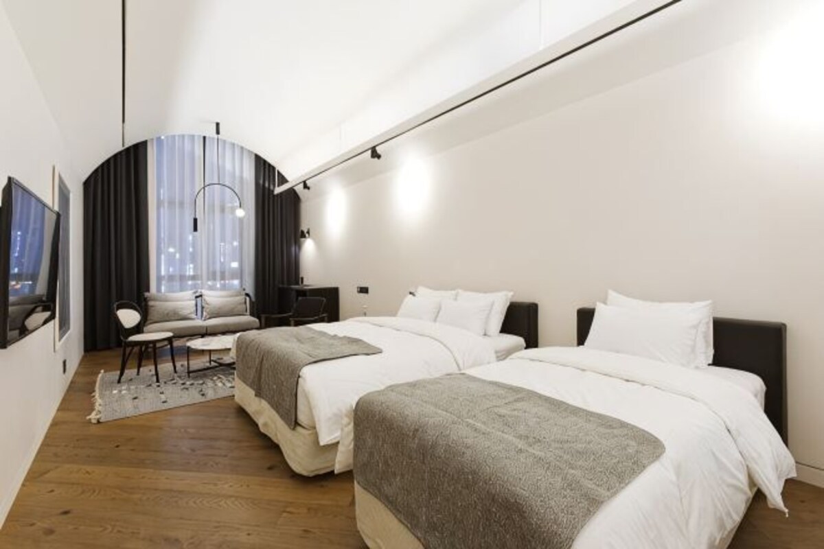 A modern hotel room with two beds, a desk, a chair, and a large window.